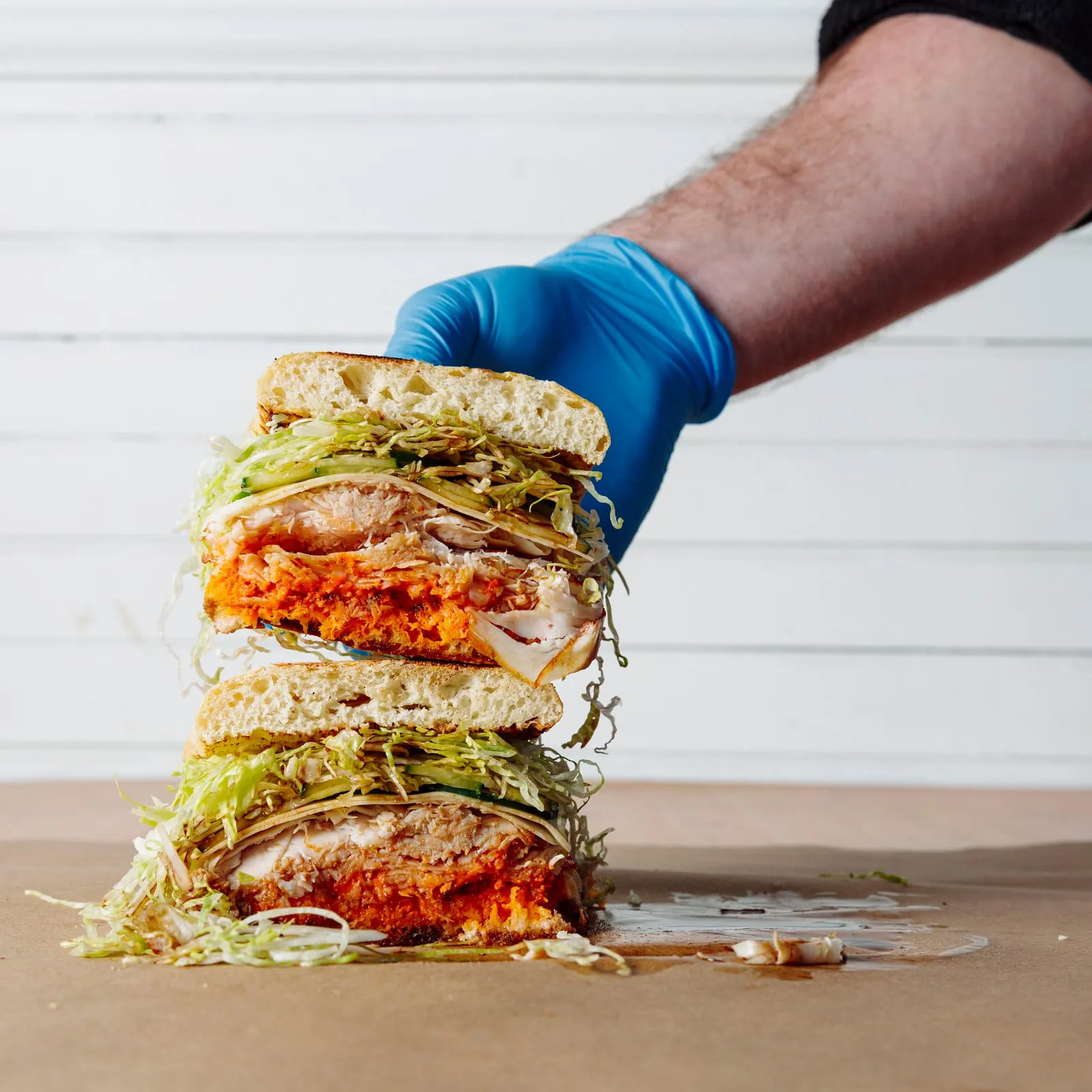 A gloved hand pressing down a tall stacked sandwich